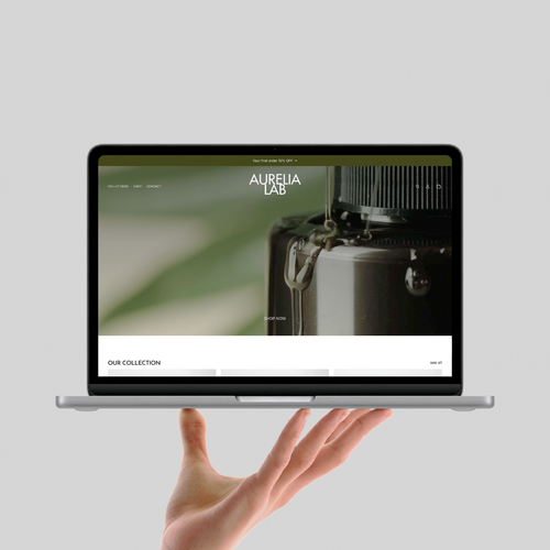 A silver laptop held up by a person's hand against a light gray background. The laptop screen displays the Aurelia Lab website with a close-up photo of a coffee maker.